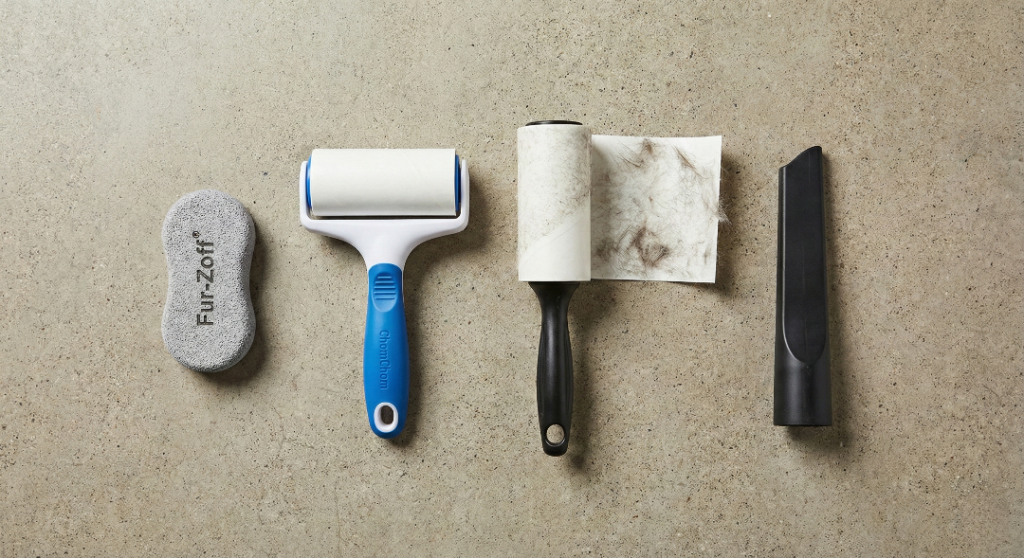 Studio product photography, flat lay style on a neutral concrete background. Four pet hair cleaning tools arranged neatly in a row